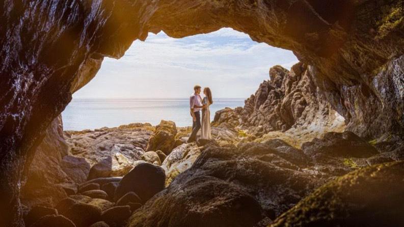 A photo showing a bride and groom facing eachother with the sea behind them, viewed from a rocky cave aperture.