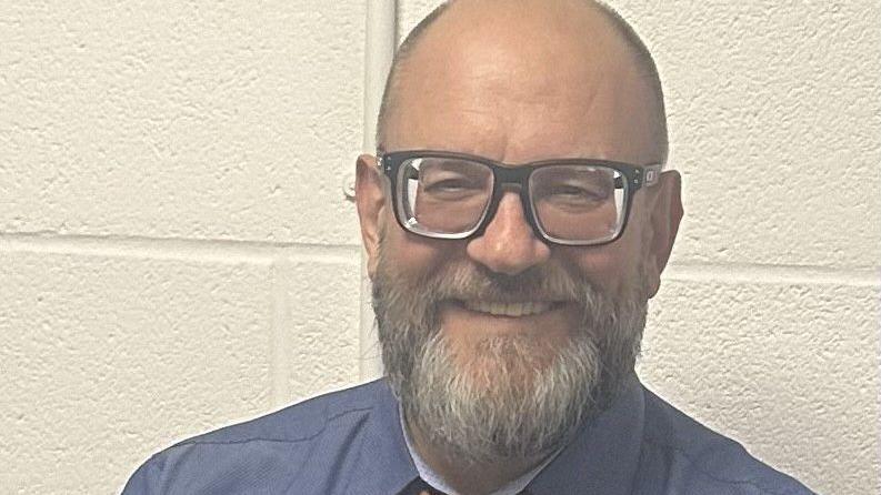Close up of Chris smiling whilst looking at the camera. He stands in front of a white bricked wall and wears a blue shirt and black glasses. He has short grey hair and a grey beard. 