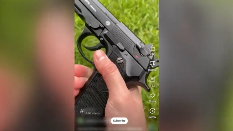 A screengrab from a TikTok video showing a hand holding a black handgun with grass in the background.