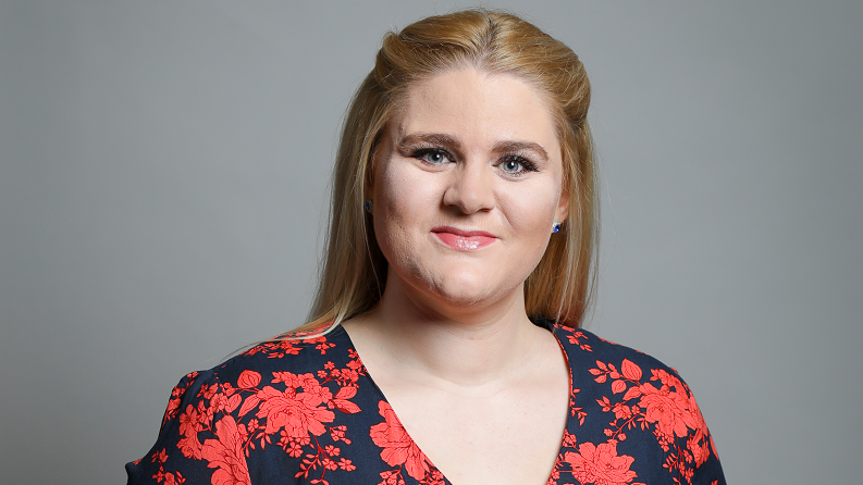 Alex Davies-Jones' official House of Commons portrait. She has long blonde hair and is wearing a black dress with a red floral print across it 