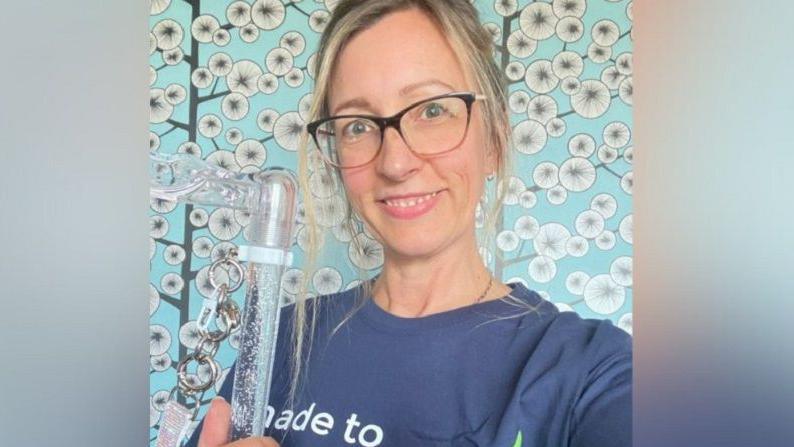 Ami Hallgarth with a blue T-shirt on and holding a clear tube. She has her blonde hair in a ponytail and is wearing glasses and is stood in front of a blue background with what appears to be wallpaper with trees that have white blossoms on them.