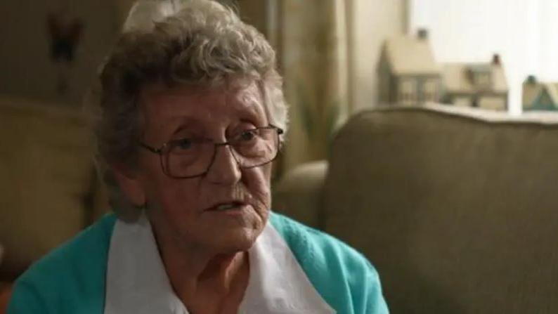 A woman with grey curly hair and glasses wearing a blue and white jumper, sat on a cream sofa in the living room of a home.