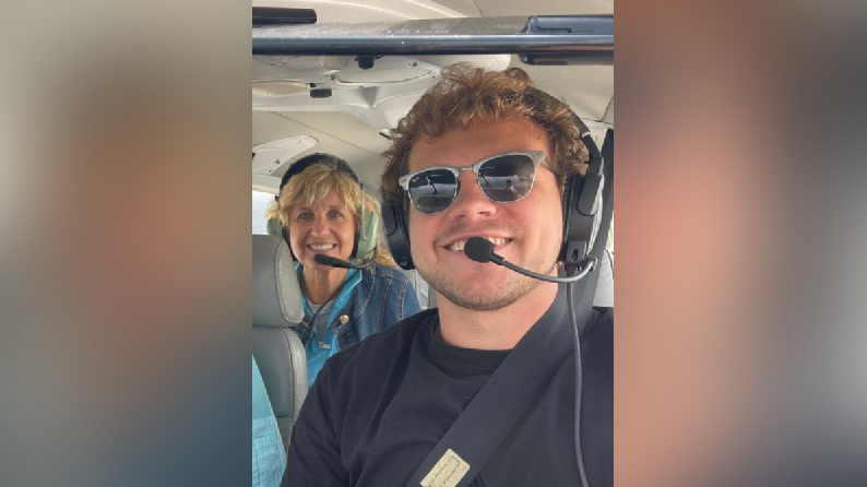 A man with curly hair, headphones and dark glasses is sitting in the cockpit of a plane. Behind him is a blonde woman in a denim jacket.