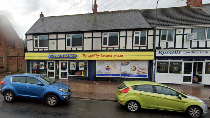 Heron Foods is in a mock tudor row of terraced houses and has a yellow shop front.  It has been raining and two cars are parked in front of the shop