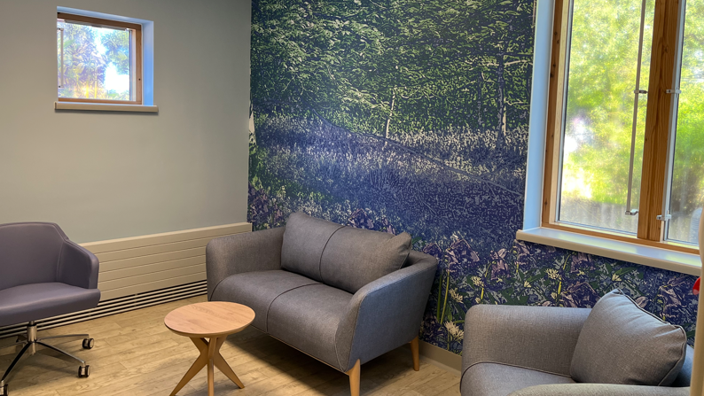 A hospital sitting area with blue and green flower wall, with lilac seats and a small round wooden table. There are also two windows behind the seating area.