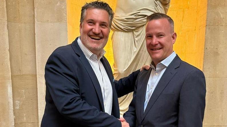 Two men wearing dark grey suits and pale shirts with no ties smile at the camera as they shake hands. They are members of the Bristol Hoteliers Association