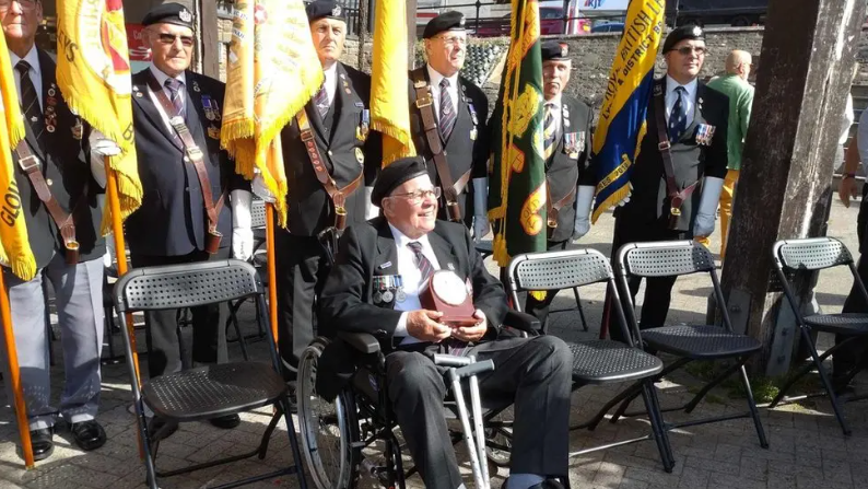 Roy Mills in a wheelchair dressed in a suit and an army cap with his medals on the lapel. Behind him six men are in similar dress, holding large yellow regimental flags.