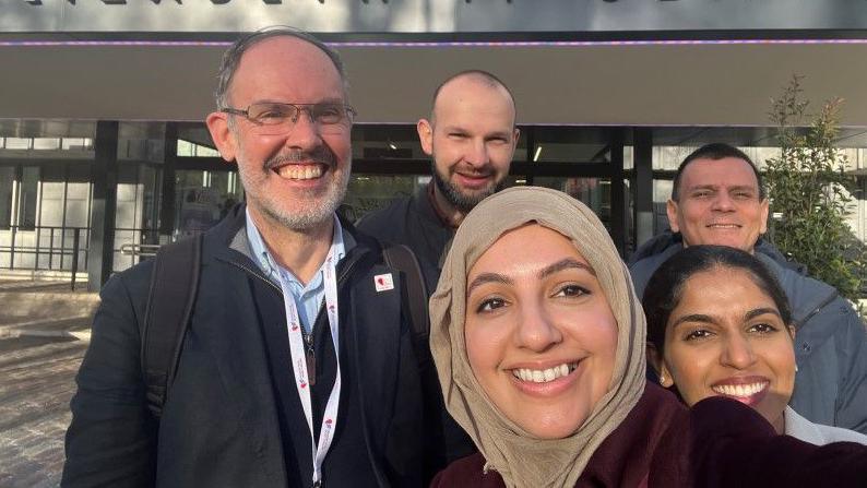 Three men and two women in front of the entrance to the building, all smiling at the camera