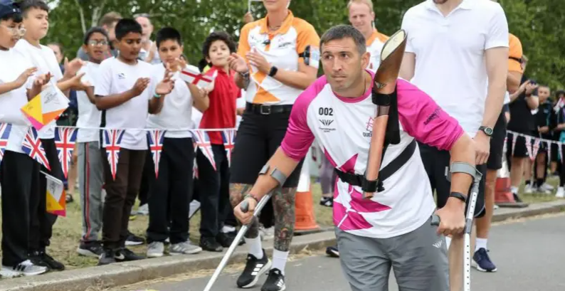 Ben Parkinson gjatë stafetës së Queen's Baton në Birmingham 2022 për të shënuar Lojën e Komonuelthit - ai mban veshur një bluzë rozë dhe të bardhë dhe përdor paterica me një shprehje të vendosur në fytyrë. Pas tij është e dukshme një turmë fëmijësh që e brohorasin ndërsa qëndron pas një kordoni të përbërë nga flamuj të vegjël të bashkimit.