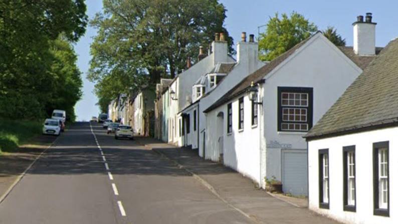 A long straight road with houses on one side and trees on the other. Some parked cars are in the distance.