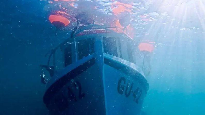 An underwater image showing a white fishing boat with black stripes and writing "GU-42". The boat is fully submerged.
