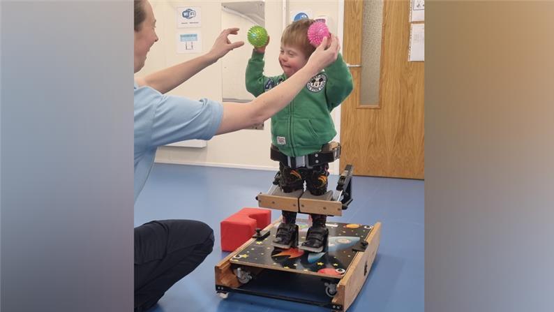 A young boy with dark blonde hair is standing in a specialised standing frame which is on wheels. He has wooden blocks in front of his knees and there is a waist strap which is undone behind him. A woman is reaching out to his hands, where he is holding a pink ball in one, and a green ball in the other, with his arms in the air.