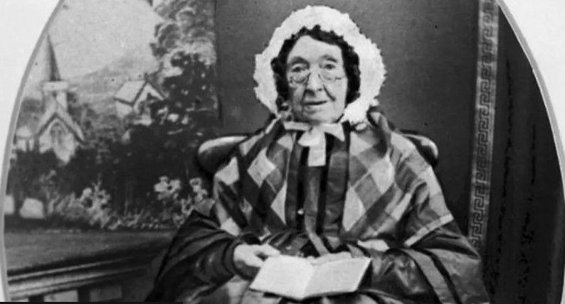 A black and white photo of an elderly lady sat on a chair. She is wearing a white bonnet on her head, a chequered scarf and long frock. She is holding an open book and looking directly at the camera. 