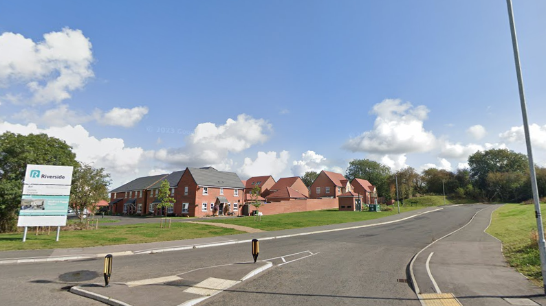 A Google Streetview looking north east from the Welford Road roundabout in Wigston. Part of a housing development surrounded by a green space and a road can be seen.