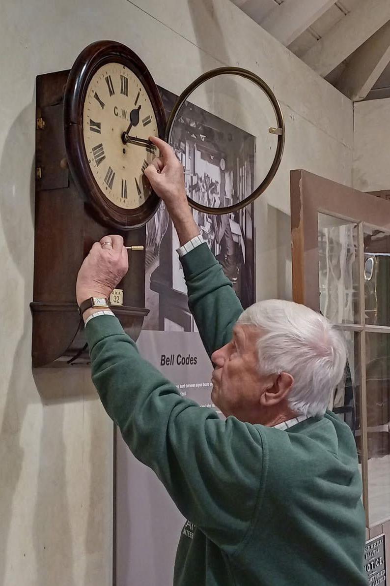 A man with white hair, wearing a green jumper, reaching up to adjust clock hands on a wall-mounted wooden clock.