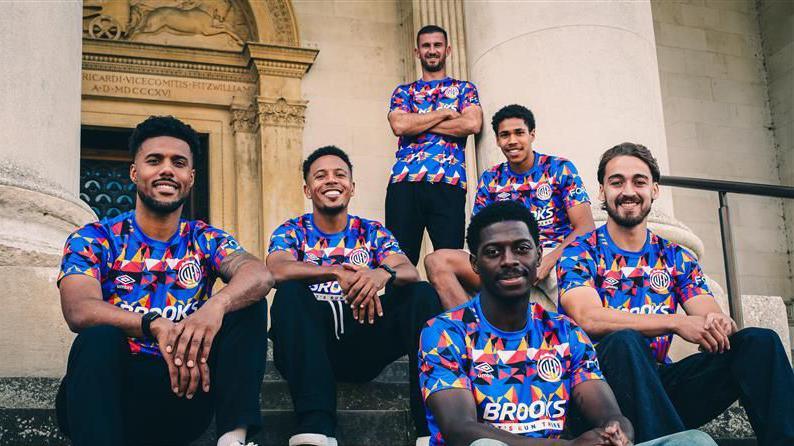 Six men are sitting on the ornate steps outside a museum. They are wearing football shirts with colourful abstract designs are on a blue background