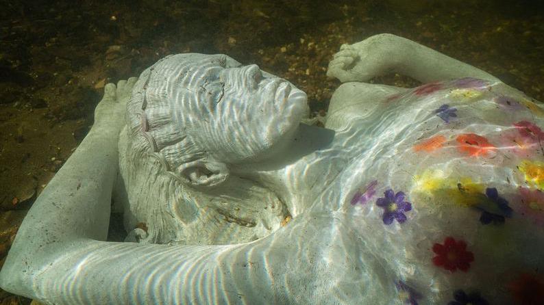 A glass statue of a woman lies underwater, she has florals painted on her bust