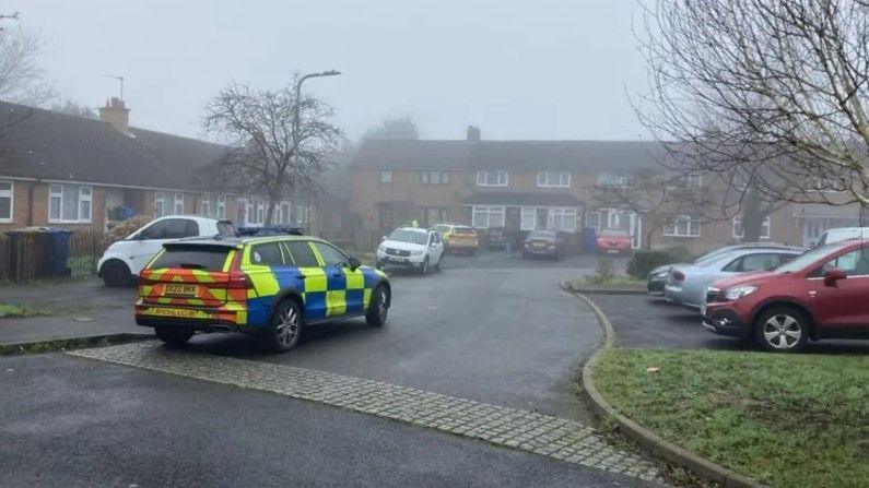 A police car is parked in a cul-de-sac. There are several houses with cars parked outside them. It is misty and grey.