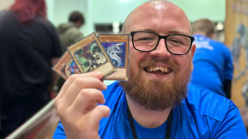 A bald man with a beard, wearing glasses, smiles as he holds up three Yu-Gi-Oh playing cards.