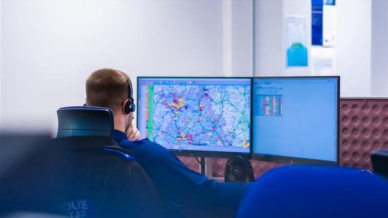 A police call handler can be seen from behind in an office. He is wearing a blue uniform and has short brown hair and a beard. He has headphones on his head and there are two computer screens in front of him, one showing a map.