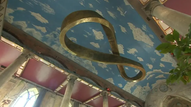 The ceiling inside the Mount Without church. It has clouds painted on to it with a large gold infinity sculpture suspended from it. The roof is supported by columns.