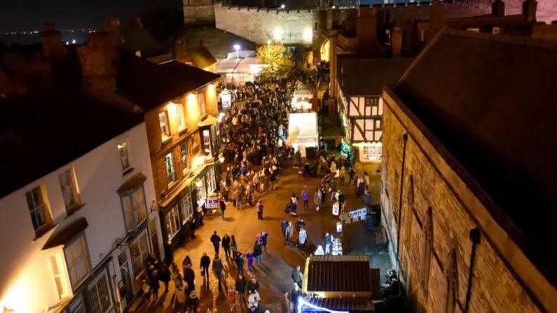 A bird's-eye view of the streets of Lincoln in the evening. There are crowds of people gathering around market stalls. The walls of Lincoln Castle are in the background