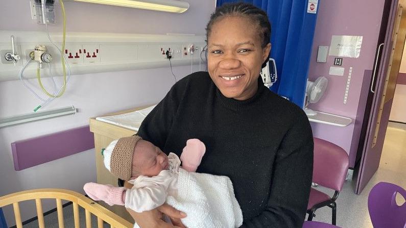 Faidat Hassan standing up holding baby Grace. They are on a maternity ward. The mother is wearing a black jumper and her baby is wrapped up warm. 