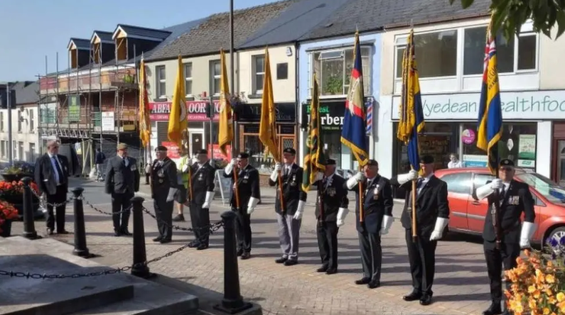 Eight men stand in a line on a town street in suits, holding a large flag each. 