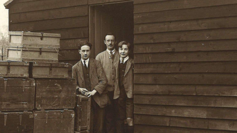 An old black & white picture showing the doorway of a large wooden hut with three men stood in it, dressed smartly in jackets and ties. There are a stack of trunks and boxes outside, presumably filled with books