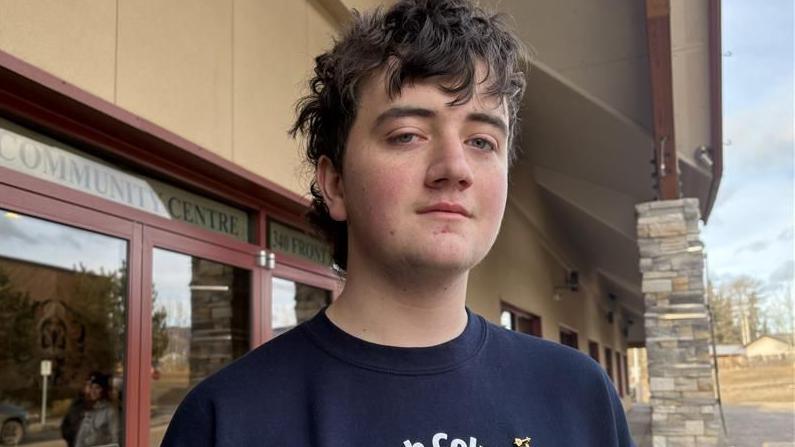 Darian Quist in a navy blue T-shirt looks at the camera with a neutral expression. Behind him is a community centre.