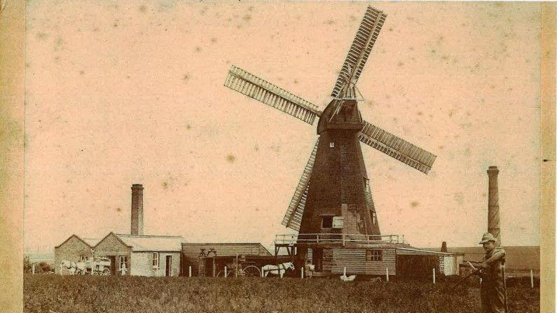 A sepia photo of a windmill next to engine rooms and sheds. There is a workmen standing near the windmill.