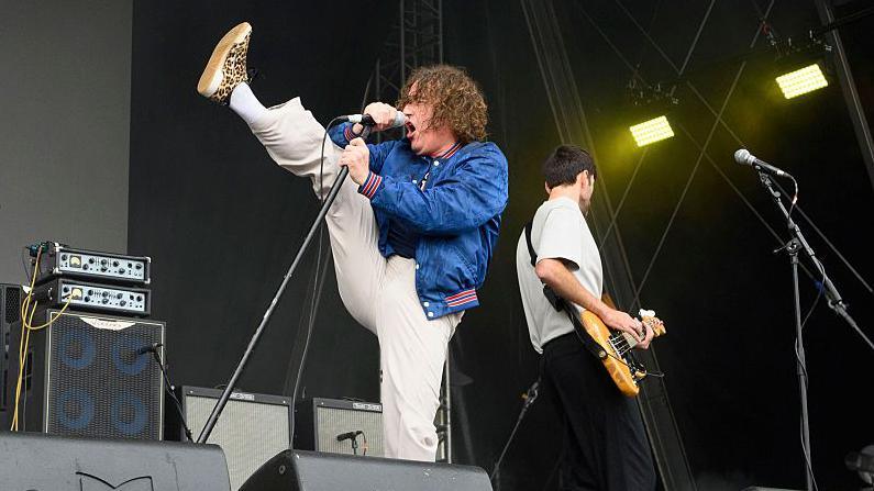 The Pigeon Detectives' Matt Bowman dancing with his right foot in the air as he sings into a microphone stand.