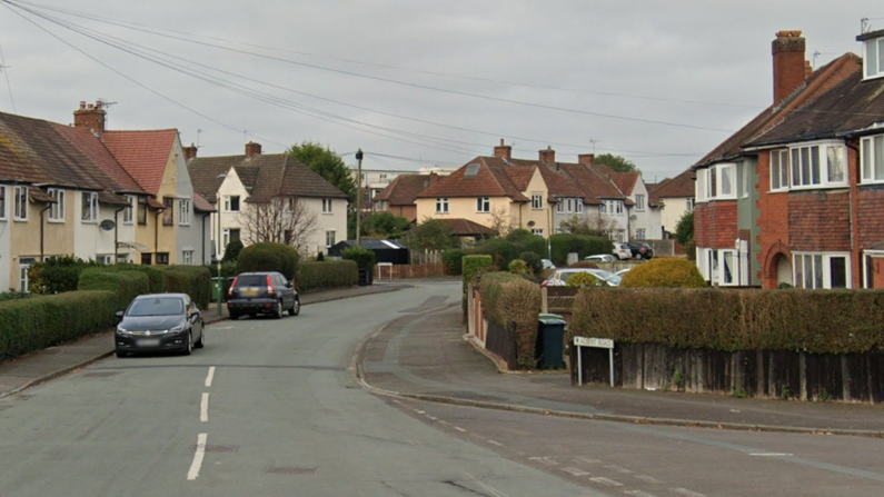 A residential street with cars parked on either side. A junction of the road can be seen heading off to the bottom right of the photo. There is a dotted white line up the centre of part of the road but it disappears further down. The houses on one side are flat-fronted with different colours to the walls including light blue and cream while the homes opposite are mainly red brick with bay windows.