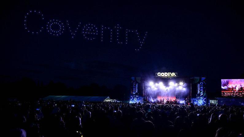 A festival stage lit up at night. The words Godiva can be seen in bright lights. In the sky, drones make up the word Coventry. Crowds of people are gathered below.