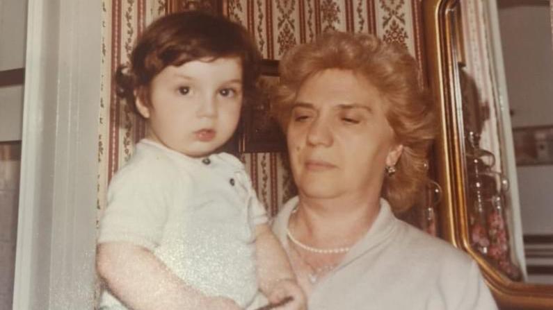 An old sepia photograph of a small boy in a white babygrow being held in his grandmother's arms. She has short, curled hair and is standing in front of a mirror.