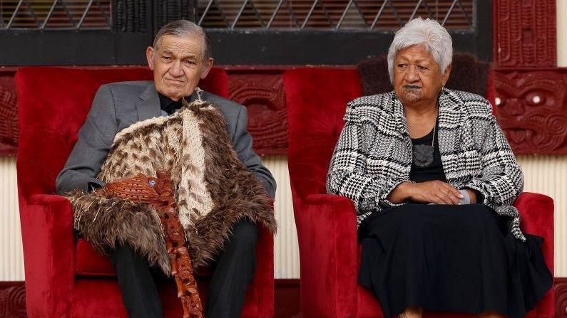 The king and his wife sit on chairs beside one another 