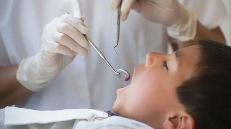 A close up of a young boy with his mouth open in a dentist's chair. A dentist is standing over him, wearing white scrubs and white latex gloves. They are using tools to examine the patient's teeth.