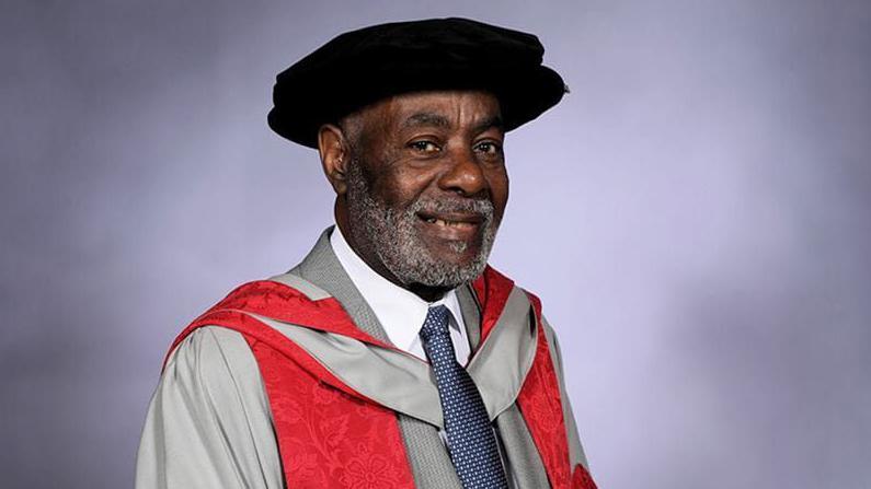 Guy Bailey's graduation photo. He is wearing grey robes with a red lapel and a black PhD cap. He is standing against a blurry purple-grey background. 