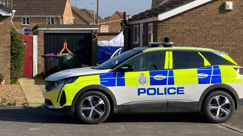 A police car is parked outside a bungalow. A police tent can be seen in the garden looking down a path at the side of the house.