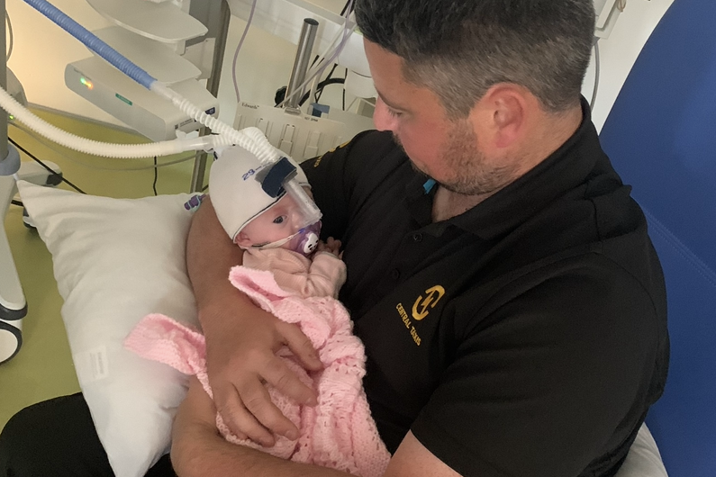 Maeva is in her dad's arms in hospital. She is in a pink blanket is wearing a hite hat. She has a ventilator on her face with a tube running upwards and out of shot.