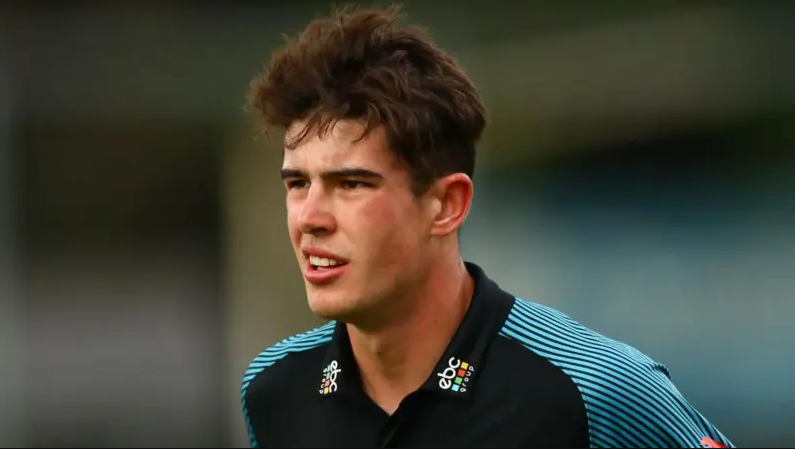 Late cricketer Josh Baker, a young man with dark curly hair, looks at something off-camera. He is wearing a black and blue cricketing top.