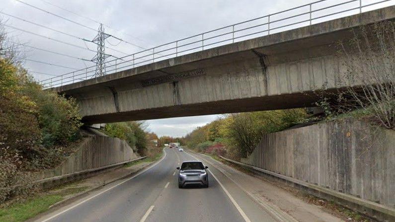 A concrete railway bridge over a modern bypass. Concrete walls support the bridge, but elsewhere, semi-mature trees line both sides of the route.