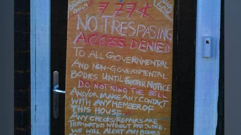 A wooden board with chalk writing on it, is attached to the front door of a property.