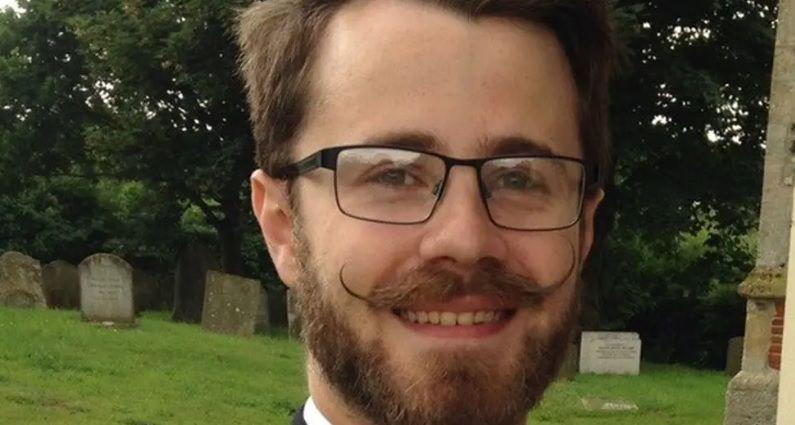 Owen Gardner wearing glasses stood outside a church. He has a handlebar moustache with turned-up ends