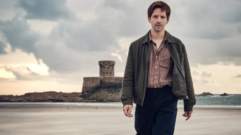 Damian Moloney as Jim Bergerac on a beach in Jersey. He is wearing a jacket and a button up shirt and black trousers. The man has brown hair and a facial hair