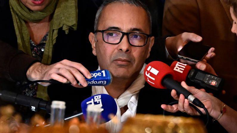 Kamel Daoud looks on pensively as he answers questions from a throng of journalists. He is sat at a table and surrounded by hands holding microphones, audio recorders and smartphones.