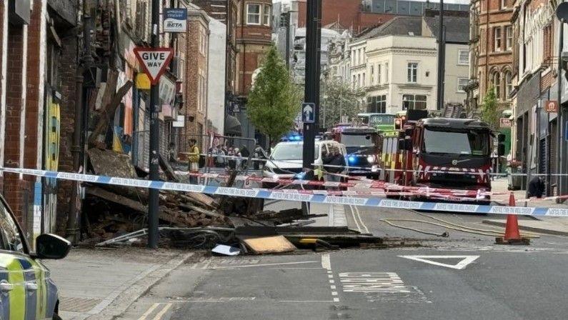 The collapsed frontage of a building lies behind a blue and white police cordon, with a police car to the left of the image and emergency vehicles, including fire engines and a police van with blue lights flashing, in the background.