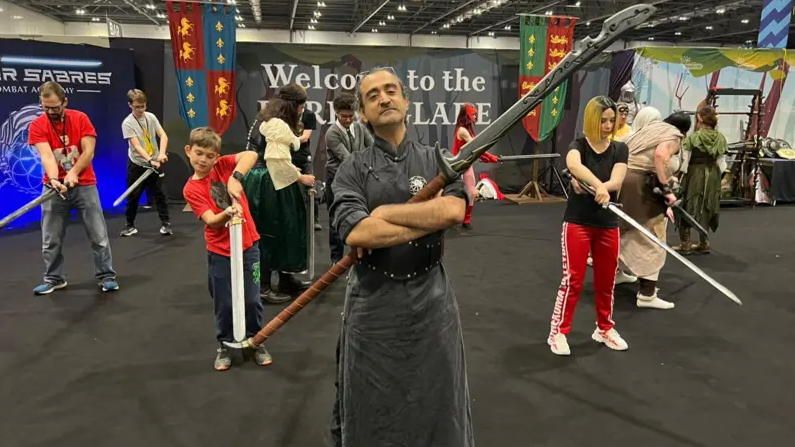 Several people wield swords behind a man in the centre, who has his arms folded over a larger sword