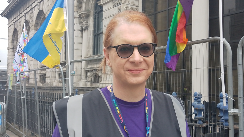 Kirsty smiling at the camera. In the background temporary fencing and pride flags  