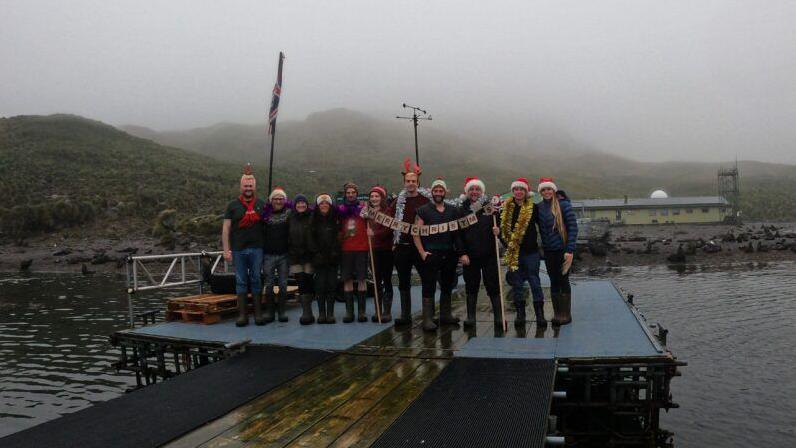 People are standing on a jetty above water. Many are wearing Santa hats and they are holding a "Merry Christmas" banner